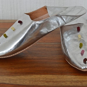 Op de afbeelding: Een paar zilveren leren slippers met een puntige neus. De slippers zijn versierd met kleine, kleurrijke, geborduurde accenten. De binnenkant van de slippers is lichtbruin.