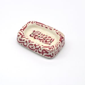 May include: A white ceramic soap dish with a red Arabic calligraphy design. The dish has a rectangular shape with a rounded oval soap well.