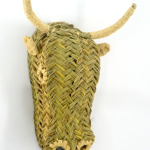 May include: A woven bull's head wall decoration made from natural materials. The head is brown and green with a woven texture. The horns are made of a light brown material.
