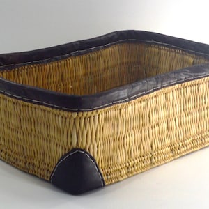 May include: A rectangular woven basket with a brown leather trim. The basket is made of natural materials and has a rustic look.