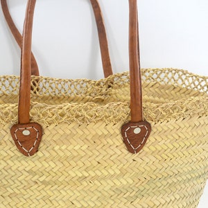 K&ouml;nnte beinhalten: Eine gro&szlig;e, naturgeflochtene Strohtasche mit braunen Ledergriffen und dekorativen N&auml;hten. Die Tasche hat einen geh&auml;kelten Besatz am oberen Rand.