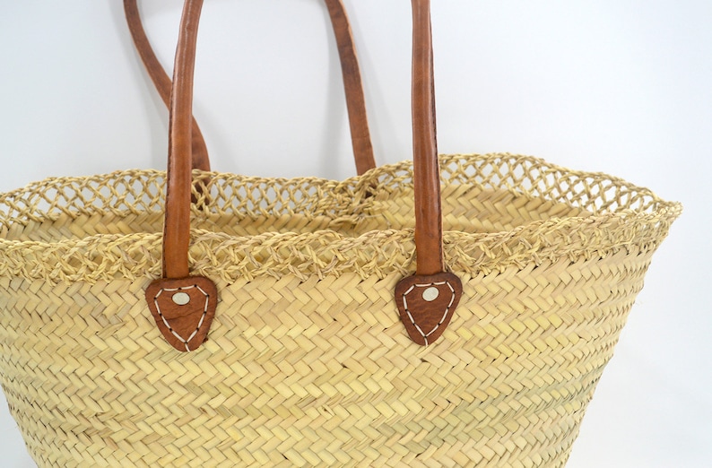K&ouml;nnte beinhalten: Eine gro&szlig;e, naturgeflochtene Strohtasche mit braunen Ledergriffen und dekorativen N&auml;hten. Die Tasche hat einen geh&auml;kelten Besatz am oberen Rand.