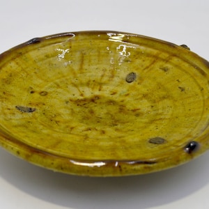 May include: A yellow ceramic dish with a slightly raised rim and a textured surface. The dish has a glossy finish and several dark brown spots around the rim.