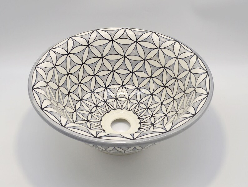 May include: Oval ceramic sink with a floral pattern in black, white, and gray. The sink has a light gray rim and a central drain hole. The design features a repeating geometric flower motif, creating a decorative and functional bathroom fixture.