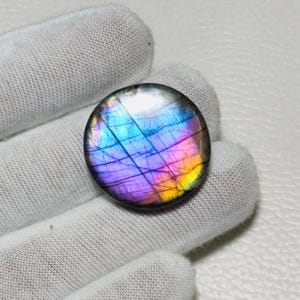 Pode incluir: Uma pedra preciosa de labradorita redonda e polida, com uma exibição vibrante de cores. A pedra apresenta uma mistura de azuis, roxos e amarelos, criando um efeito iridescente. A gema é segurada por uma mão enluvada.
