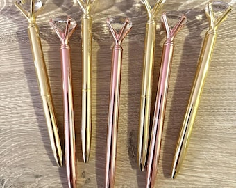 May include: A collection of stylish pens featuring diamond-shaped tops. The pens are in gold and rose gold hues, displayed on a wooden surface. The pens have a sleek, metallic design, with the diamond-shaped tops adding a touch of luxury and sophistication.