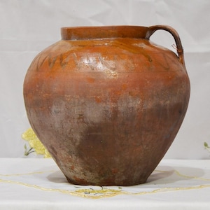 Puede incluir: Una jarra grande de cerámica de color terracota, con cuerpo redondeado y asa. La jarra tiene una boca ancha y una superficie ligeramente texturizada, con patrones decorativos descoloridos cerca de la parte superior. La apariencia general sugiere edad y encanto rústico.