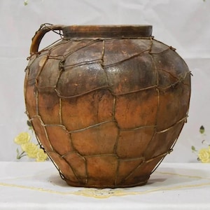 May include: A large, antique-style earthenware jug with a handle. The jug is a warm brown color, with a wire mesh wrapped around it. The jug has a rustic, aged appearance, suggesting it is an old piece. The jug is likely used for storage or decoration.