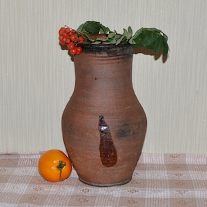 Op de afbeelding: Een bruine kleipot met een brede opening en een rond lichaam. De pot is versierd met een bruine vlek en heeft een klein takje met rode bessen erop.