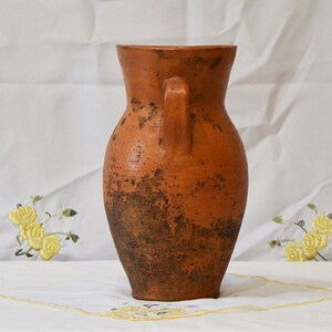 Op de afbeelding: Een terracotta vaas met een bruine glazuur en een handvat aan &eacute;&eacute;n kant. De vaas heeft een rustieke, verweerde uitstraling.