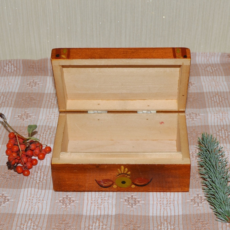 May include: A wooden box with a hinged lid, painted in shades of brown and yellow. The box has a floral design painted on the front and is open to reveal the empty interior.
