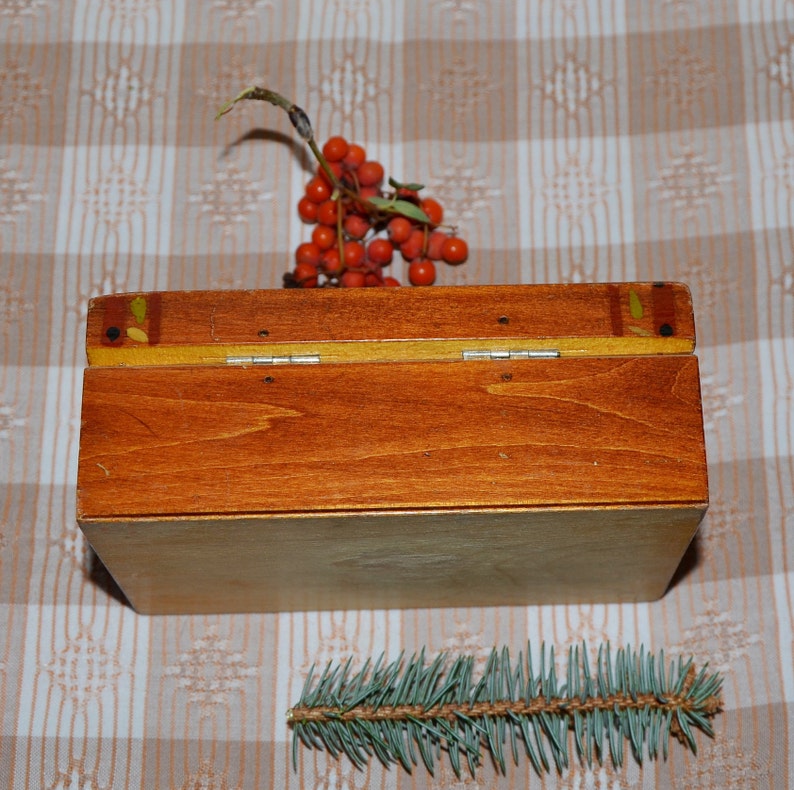 May include: A wooden box with a hinged lid, painted in a warm brown color with yellow accents. The box is closed and sitting on a checkered tablecloth. A sprig of red berries and a small evergreen sprig are also visible.