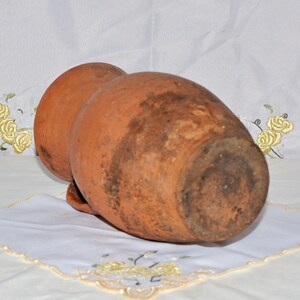 Op de afbeelding: Een terracotta kruik met een rond lichaam en een smalle hals. De kruik heeft een ruw, getextureerd oppervlak en is licht verweerd.