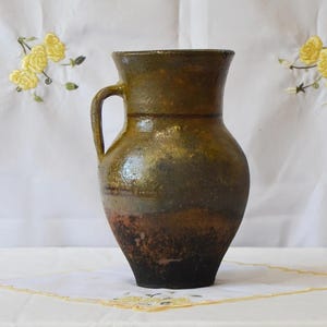 Puede incluir: Jarra de cerámica rústica y artesanal con asa. La jarra presenta un degradado de colores, que va del marrón oscuro en la base al verde oliva y dorado en la parte superior. La jarra probablemente se utiliza con fines decorativos.