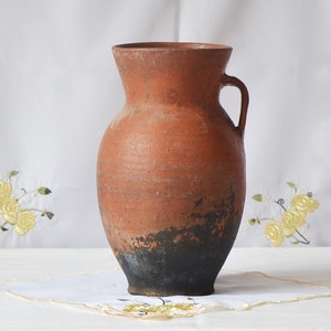 May include: A terracotta vase with a dark brown glaze, featuring a rounded body and a single handle. The vase is sitting on a white tablecloth.