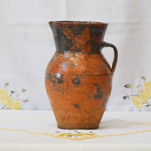 Op de afbeelding: Een rustieke keramische kan met een handvat. De kan heeft een warme, oranje-bruine basiskleur met donkerbruine en zwarte gevlekte accenten. De rand en de binnenkant zijn donkerder bruin. Dit vintage stuk is waarschijnlijk voor het serveren van water of dranken.