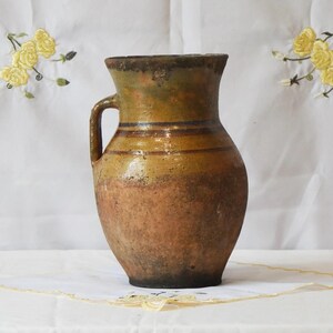 May include: A vintage pottery jug with a handle, featuring a rounded body and a flared rim. The colour scheme includes shades of brown, tan, and olive green, with horizontal bands adding detail. The jug is likely handmade and suitable for decorative use.