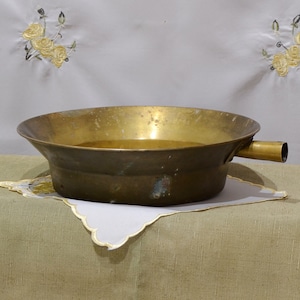 May include: A brass bowl with a spout on the side, sitting on a white doily on a brown linen tablecloth.
