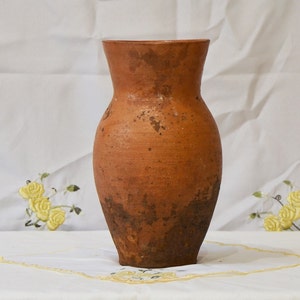 Op de afbeelding: Een terracotta vaas met een rustieke, aardse bruine kleur en een licht onregelmatig oppervlak. De vaas heeft een brede basis en een smalle hals, wat hem een klassieke, traditionele uitstraling geeft.
