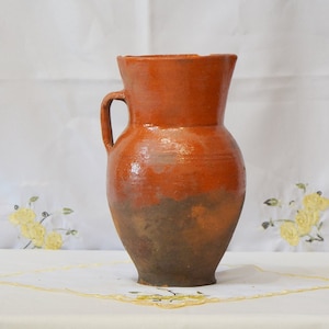Op de afbeelding: Een terracotta kan met een handvat, geglazuurd in een roodbruine kleur. De kan heeft een licht onregelmatig oppervlak en een donkerbruine kleur aan de onderkant.