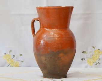 Op de afbeelding: Een terracotta kan met een handvat, geglazuurd in een roodbruine kleur. De kan heeft een licht onregelmatig oppervlak en een donkerbruine kleur aan de onderkant.