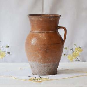 May include: A brown clay pitcher with a handle. The pitcher has a white glaze on the bottom and a brown glaze on the top. The pitcher is sitting on a white tablecloth.