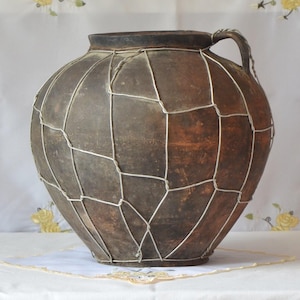 Puede incluir: Una jarra grande de cerámica marrón antigua con asa, envuelta en una malla de alambre plateado. La jarra tiene un cuerpo redondeado y un borde oscuro. La malla de alambre tiene un patrón geométrico.
