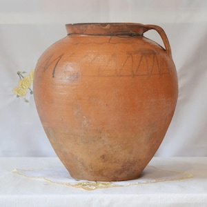 May include: A large, antique terracotta jar with a handle. The jar has a wide, rounded body and a narrow neck. It features faded black markings and a decorative line near the top. The jar is a warm, earthy brown color.