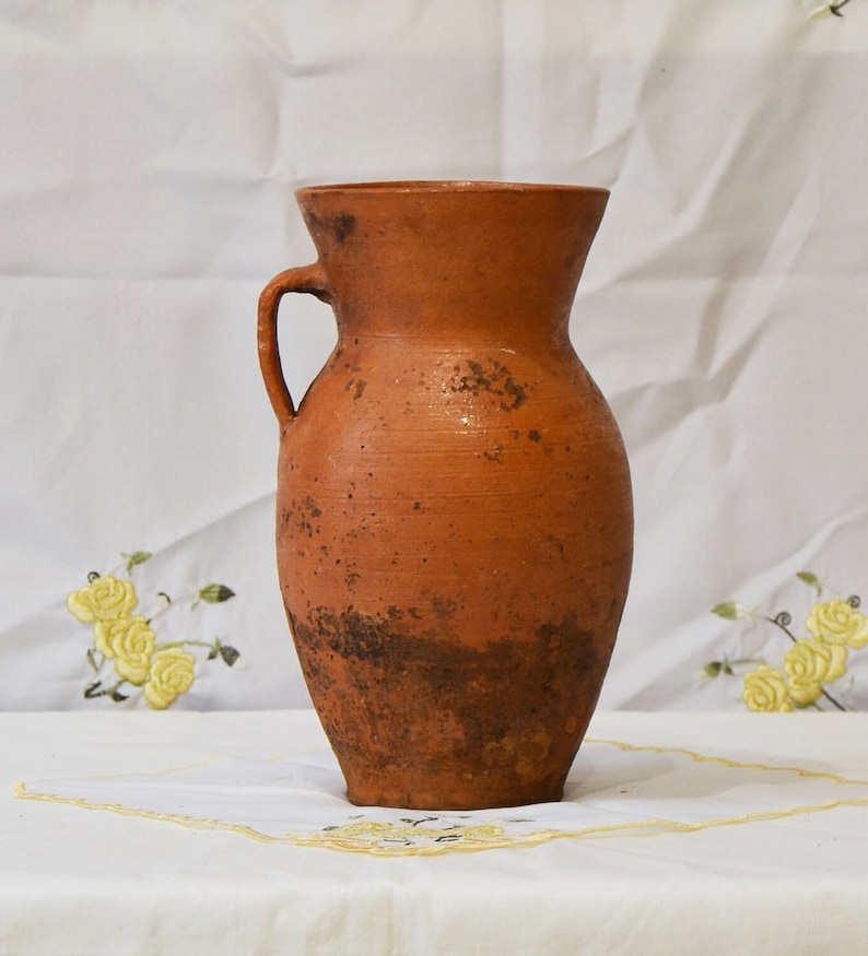 Op de afbeelding: Een terracotta klei kan met een handvat. De kan heeft een rustieke, verweerde uitstraling met bruine en oranje tinten.