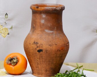 Op de afbeelding: Een rustieke bruine kleipot met een brede opening en een rond lichaam. De pot staat op een wit tafelkleed met een kaki-vrucht ernaast.