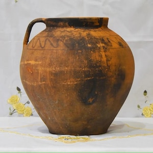 Könnte beinhalten: Ein großer, runder Krug aus Steingut mit Henkel. Der Krug hat eine warme braune Farbe mit dunkelbraunen und schwarzen Akzenten. Ein dekoratives Wellenlinienmuster befindet sich oben. Der Krug steht auf einer weißen Oberfläche mit gelben Blumenakzenten.