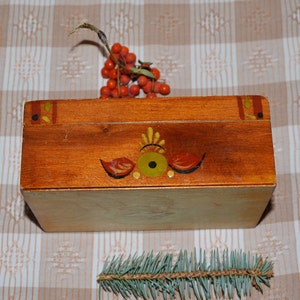 May include: A wooden box with a painted design of a stylized eye and floral motifs. The box is brown with yellow, red, and black accents. The box is closed and sitting on a checkered tablecloth.