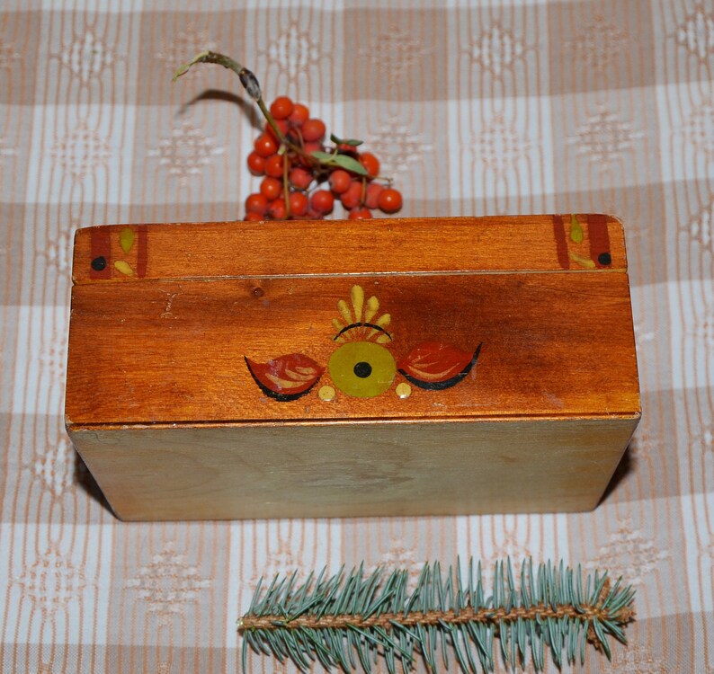 May include: A wooden box with a painted design of a stylized eye and floral motifs. The box is brown with yellow, red, and black accents. The box is closed and sitting on a checkered tablecloth.