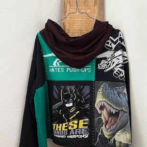May include: Black patchwork hoodie with Batman, a T-Rex, and a spaceship graphic. The hoodie has a green panel with the text "Hates Push-Ups" and the phrase "These Hands Are Mastered Weapons".