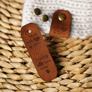 Può includere: Due etichette in pelle marrone con il testo "Made with love by Sophia" e un design a forma di cuore. Le etichette sono su una superficie di vimini intrecciato.