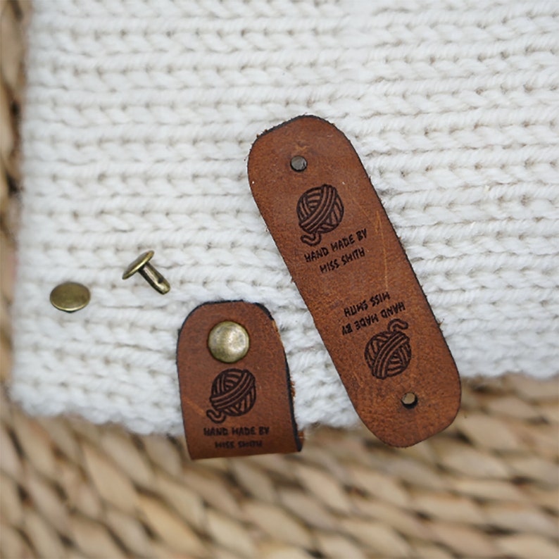 Op de afbeelding: Twee bruine leren labels met een garenbal-ontwerp en de tekst "HAND MADE BY MISS SMITH" en "HAND MADE BY MISS CHINN".