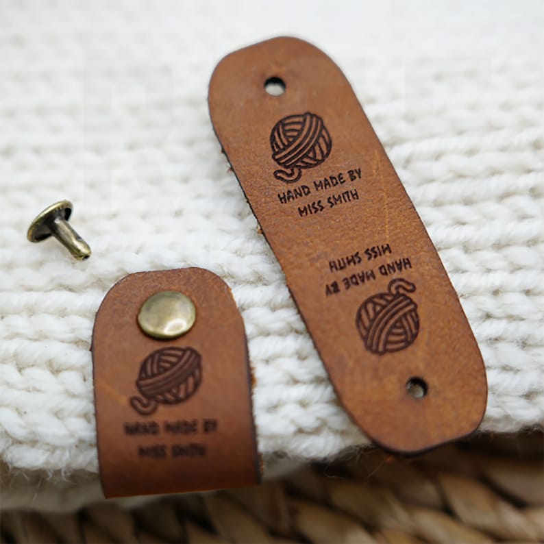 Op de afbeelding: Twee bruine leren labels met een garenbal-ontwerp en de tekst "HAND MADE BY MISS SMITH" op het ene label en "HAND MADE BY MISS SMITH" op het andere label.