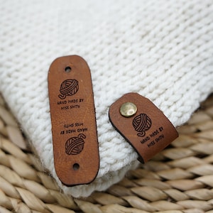 Op de afbeelding: Twee bruine leren labels met een garenbal-ontwerp en de tekst "HAND MADE BY MISS SHITH" op een witte gebreide stoffen achtergrond.