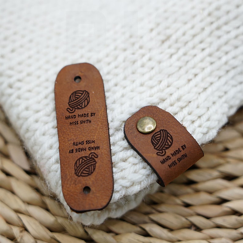 Op de afbeelding: Twee bruine leren labels met een garenbal-ontwerp en de tekst "HAND MADE BY MISS SHITH" op een witte gebreide stoffen achtergrond.