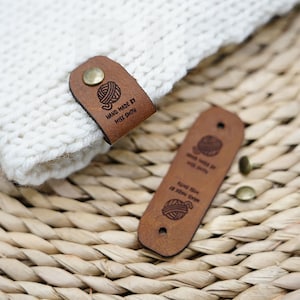 Op de afbeelding: Twee bruine leren labels met een bol garen en de tekst "HAND MADE BY MISS SMITH" erop gegraveerd. De labels liggen op een geweven rieten oppervlak.