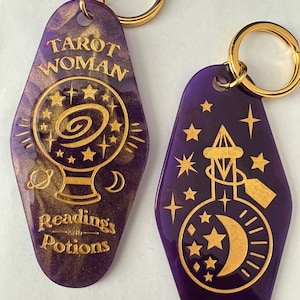 K&ouml;nnte beinhalten: Zwei lila und goldene Schl&uuml;sselanh&auml;nger mit einem goldenen Ring. Der Schl&uuml;sselanh&auml;nger links hat ein goldenes Design einer Kristallkugel mit Sternen und dem Text "Tarot Woman" und "Readings and Potions". Der Schl&uuml;sselanh&auml;nger rechts hat ein goldenes Design einer Flasche mit einem Mond und Sternen.