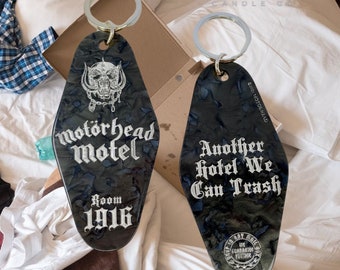 May include: Two black and silver keychains with the text "Motörhead Motel" and "Another Hotel We Can Trash". The keychains feature a skull design and the room number 1916. The keychains are on a bed.