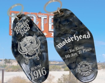 May include: Two black and silver keychains shaped like motel room keys. One keychain features the Motörhead logo and the text "You Know We Aim To Please". The other keychain features the text "The Bomber B&B" and a silver skull with horns. Both keychains have a silver ring for attaching to keys.