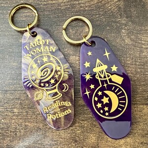 K&ouml;nnte beinhalten: Zwei lilafarbene Schl&uuml;sselanh&auml;nger mit goldenen Akzenten. Ein Schl&uuml;sselanh&auml;nger zeigt eine Kristallkugel mit Sternen und dem Text "Tarot Woman with Readings & Potions". Der andere Schl&uuml;sselanh&auml;nger zeigt eine Flasche mit einem Halbmond und Sternen.
