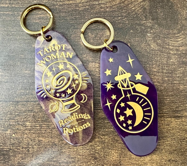 K&ouml;nnte beinhalten: Zwei lilafarbene Schl&uuml;sselanh&auml;nger mit goldenen Akzenten. Ein Schl&uuml;sselanh&auml;nger zeigt eine Kristallkugel mit Sternen und dem Text "Tarot Woman with Readings & Potions". Der andere Schl&uuml;sselanh&auml;nger zeigt eine Flasche mit einem Halbmond und Sternen.