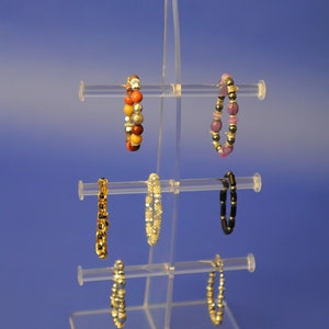 May include: A clear acrylic jewellery stand with six arms for displaying bracelets. The stand has a curved base and is designed to hold bracelets of various sizes and styles. The stand is displaying a variety of bracelets, including beaded bracelets, chain bracelets, and charm bracelets.
