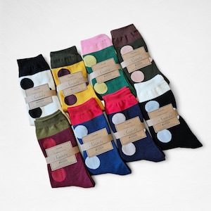 May include: A selection of colourful socks featuring large polka dots. The socks are in various colours, including black, white, yellow, green, red, and blue. Each pair is wrapped with a brown paper band that reads "Saints".