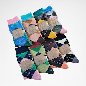 Peut inclure: Une collection de chaussettes à motifs argyle de différentes couleurs, dont le bleu, le vert, le rose et le marron. Chaque paire est enveloppée d'une bande de papier marron avec l'inscription "Saints". Les chaussettes sont en coton.