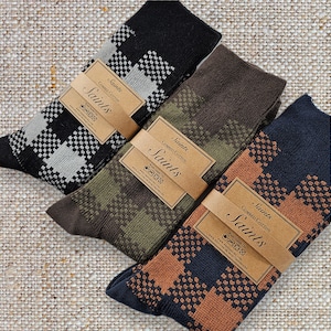 Könnte beinhalten: Drei Paar Socken mit verschiedenen geometrischen Mustern. Die Socken sind schwarz, braun und marineblau mit weißen, grünen und orangefarbenen Akzenten. Die Socken sind aus gekämmter Baumwolle und tragen den Markennamen "Saints" auf der Verpackung.