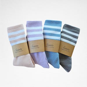 Könnte beinhalten: Vier Paar gestreifte Socken in verschiedenen Farben: Rosa, Lavendel, Blau und Grau. Jede Socke hat ein braunes Etikett mit dem Wort "Saints" und dem Text "Complete Your Style".